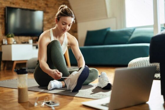 A woman getting ready to exercise at home, sitting in her living room on a yoga mat, putting on running shoes and using a laptop for blog post apps that pay you to exercise