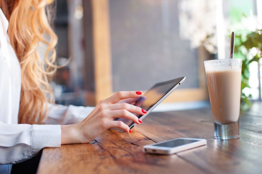 A woman working remotely from a cafe, holding a tablet while drinking coffee.