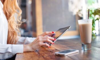 A woman working remotely from a cafe, holding a tablet while drinking coffee.