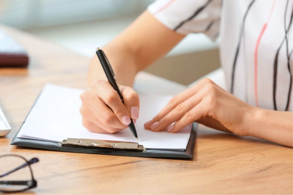 Una mujer sentada en un escritorio, escribiendo notas en un portapapeles.