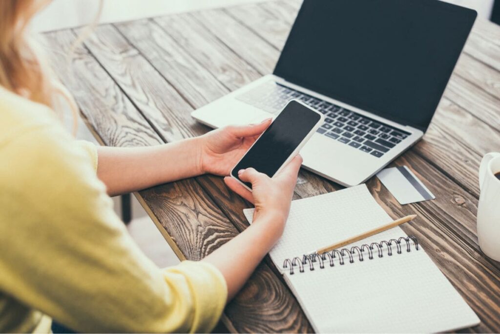 A woman working remotely from a cafe, using her phone while working on a laptop.