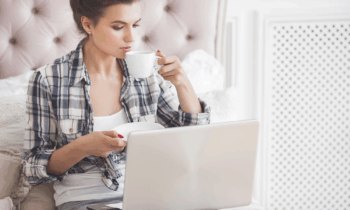 A casual young woman working on her laptop drinking coffee in her bedroom completing short task jobs