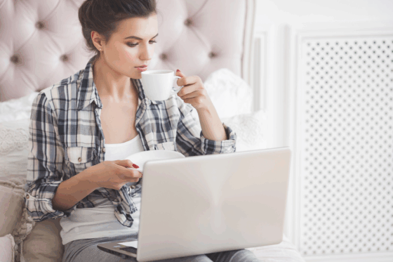 A casual young woman working on her laptop drinking coffee in her bedroom completing short task jobs