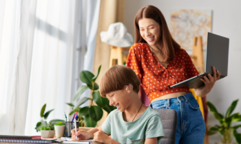 A mom standing with her laptop in hand looking over her son's shoulder while he colors for blog post summer jobs for moms
