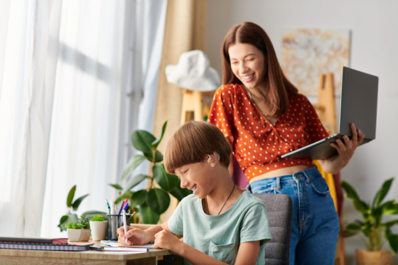 A mom standing with her laptop in hand looking over her son's shoulder while he colors for blog post summer jobs for moms