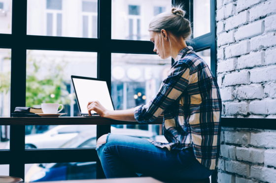 A side view of a young woman reading a User Interviews Review on her laptop in a cafe
