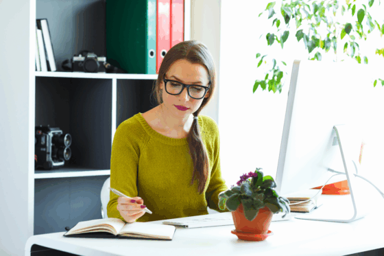 A woman wearing glasses is sitting at her desk looking at her notebook and list of non-phone work from home jobs
