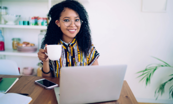 A woman with long curly hair and glasses drinking a cup of coffee while looking for ways to make money online