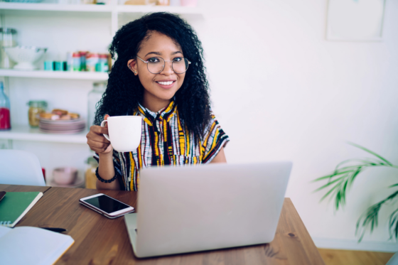 A woman with long curly hair and glasses drinking a cup of coffee while looking for ways to make money online