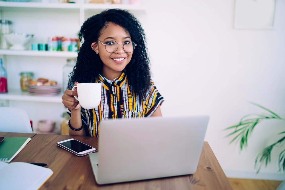 A woman with long curly hair and glasses drinking a cup of coffee while looking for ways to make money online