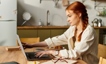 A woman with red hair in a braid working on her laptop in her kitchen looking for virtual jobs