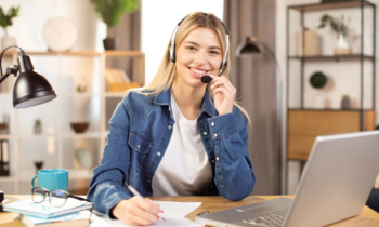 A young blond woman wearing a headset and taking notes while she provides home-based customer service support