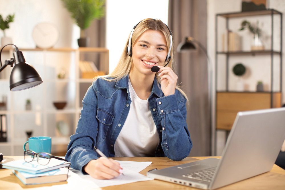 A young blond woman wearing a headset and taking notes while she provides home-based customer service support