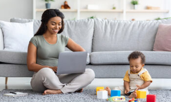 A young mom working on her laptop while her baby plays with toys next to her for work from home summer jobs