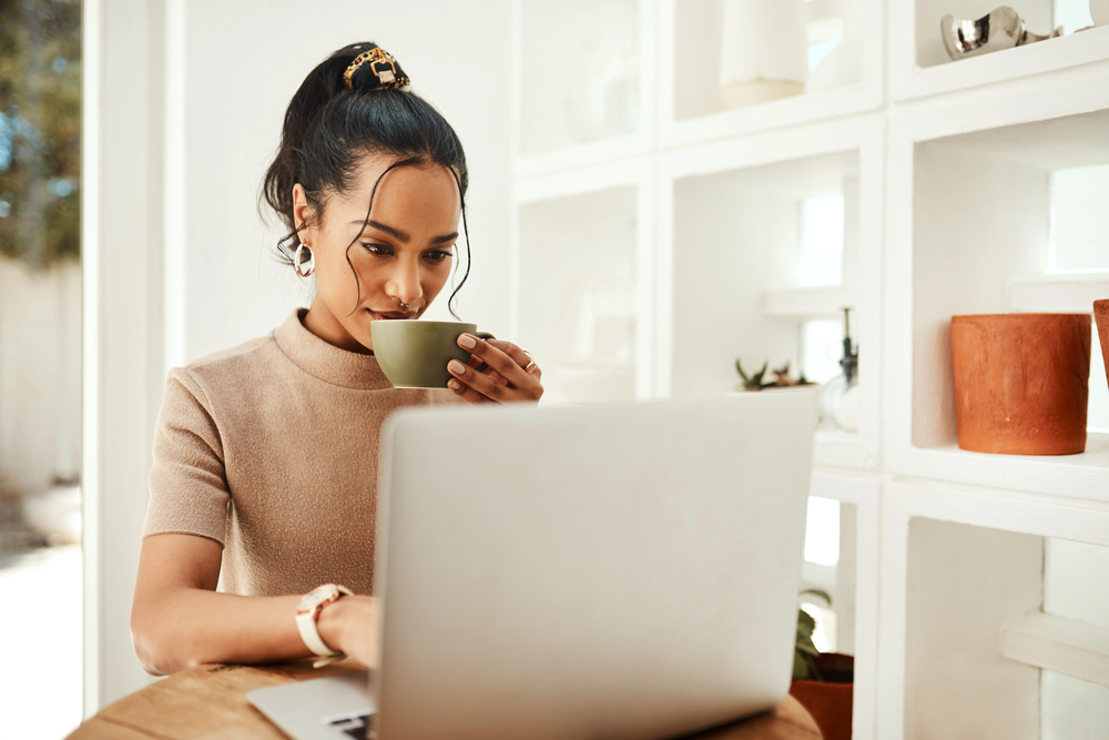 A young woman drinking coffee and reading a FlexJobs review on her laptop