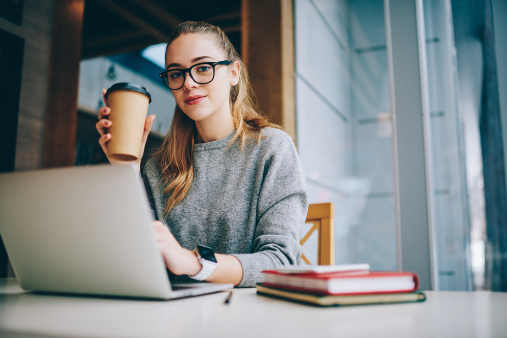 A young woman wearing glasses and drinking coffee looking for work at home jobs with employee benefits on her laptop