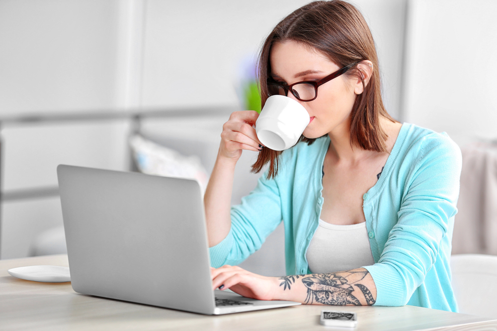 A young woman wearing glasses drinking coffee searching for remote test scoring jobs on her laptop