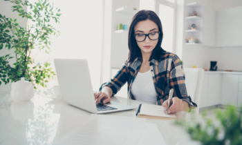 A young woman with long hair and glasses working on her laptop in her kitchen for blog post BELAY Solutions Review