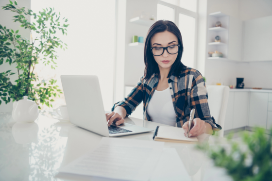 A young woman with long hair and glasses working on her laptop in her kitchen for blog post BELAY Solutions Review