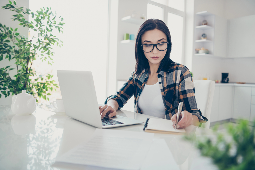 A young woman with long hair and glasses working on her laptop in her kitchen for blog post BELAY Solutions Review