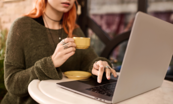 A young woman with long red hair is drinking coffee and looking at her laptop searching for remote jobs that pay weekly