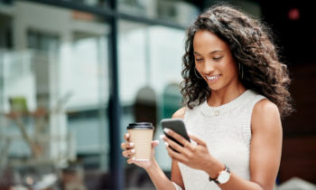 A young woman with long wavy hair is drinking coffee and reading an article on her phone titled 14 trusted money-making websites