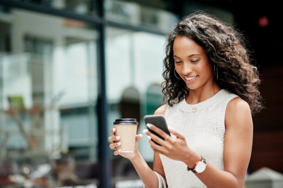 A young woman with long wavy hair is drinking coffee and reading an article on her phone titled 14 trusted money-making websites