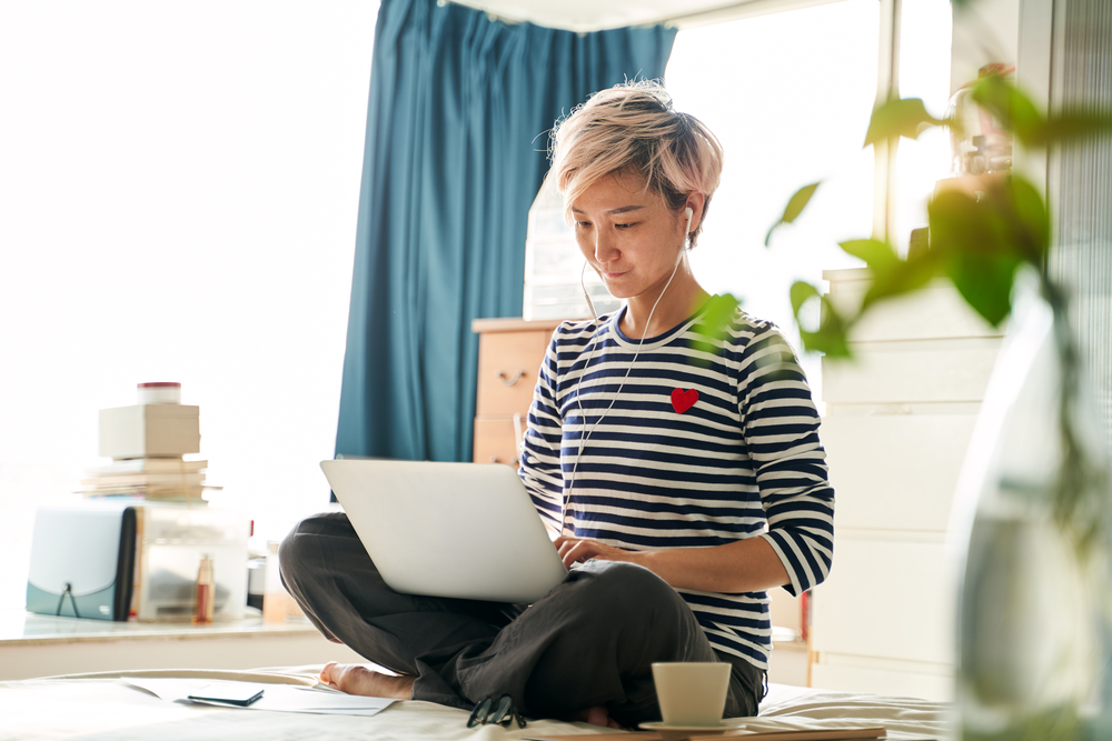 Beautiful Asian woman sitting on bed & working from home on laptop