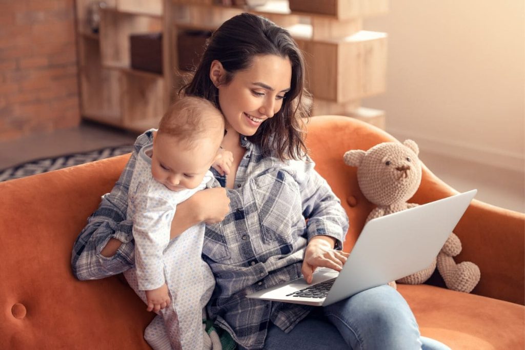 Stay at home mom working at home on sofa, while holding her baby.