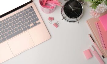 Overhead view of a remote worker's desk, with a laptop, pencils, notebooks, and clock.
