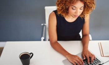 Overhead view of a woman working at her home office desk, drinking coffee and typing on a keyboard for blog post earn what you deserve.