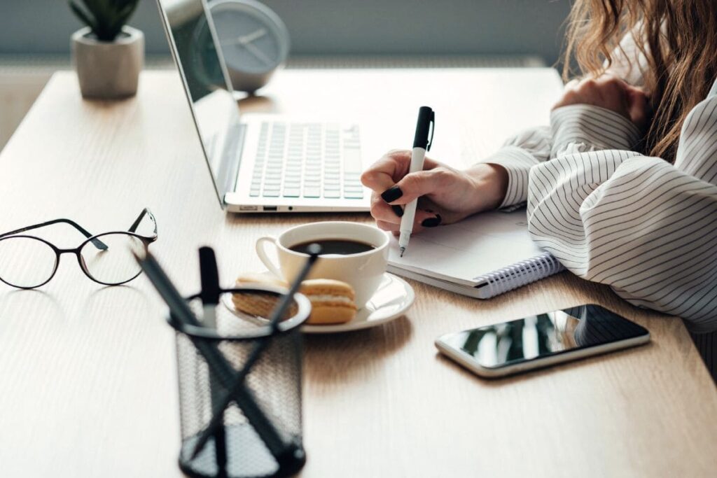 Woman sitting at desk, working on laptop and writing on notebook.
