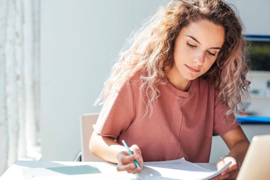 Woman sitting at desk, working on laptop and writing on notebook.