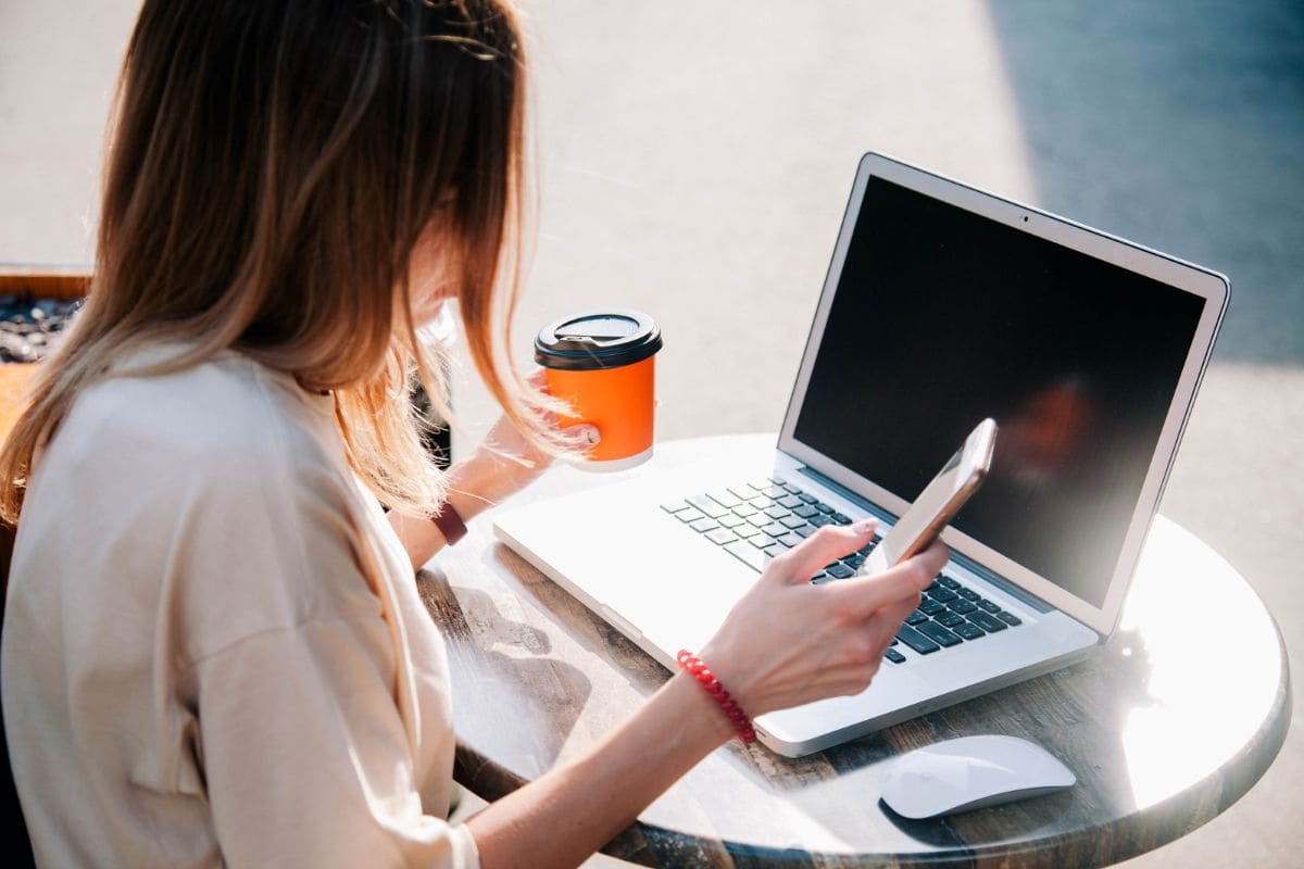 A woman sitting at an outdoor table at a cafe, using a smartphone and laptop, while drinking coffee for the blog post freecash review