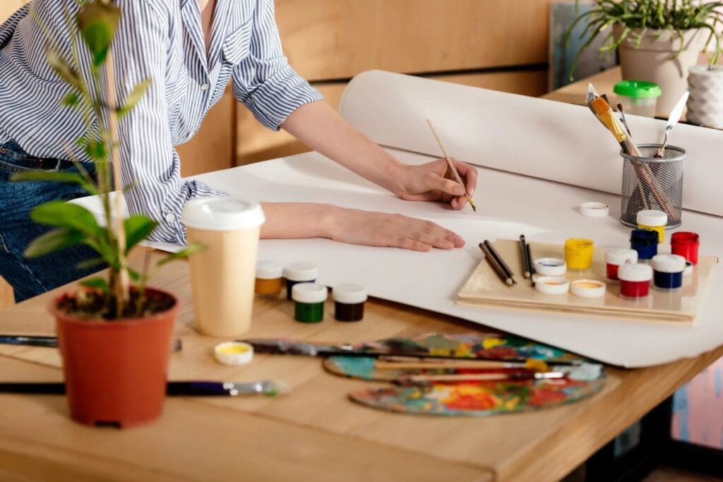 A closeup view of an artist's desk: a woman is using paints to color a large canvas.