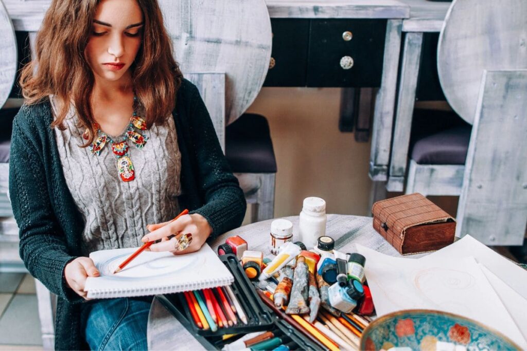 A woman sitting outdoors at a table and using colored pencils to draw in a large sketchbook.