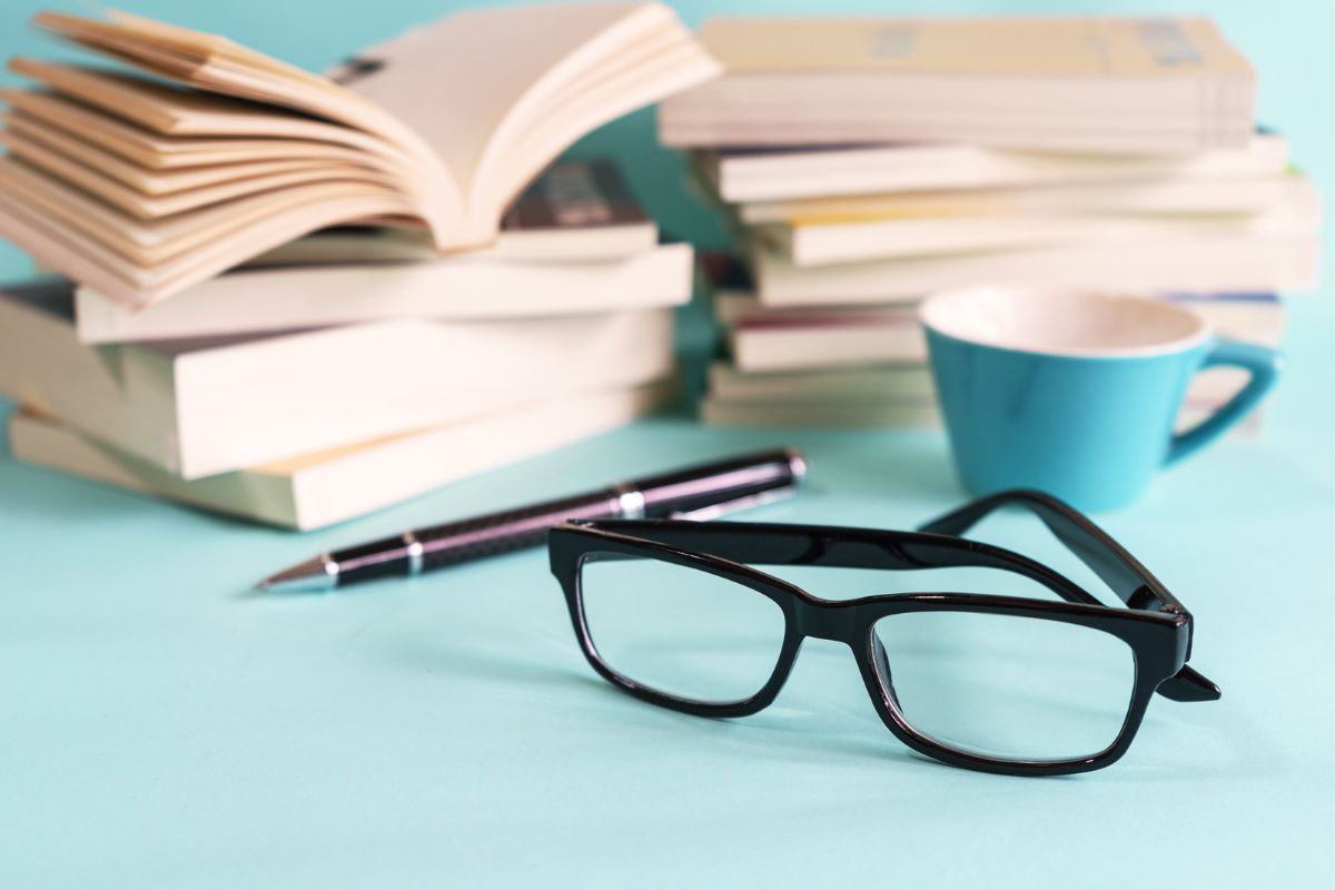 A stack of books on a desk next to a pair of glasses and a cup of coffee.