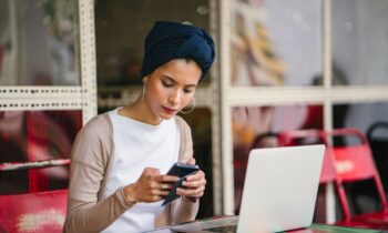 A content creator working remotely outside at a cafe, using a laptop while looking at her phone.