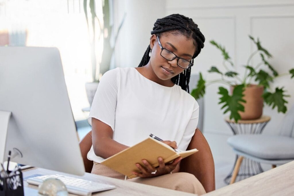 A woman working at her home office desk, using a computer and writing in a notebook.