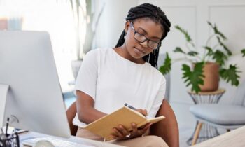 A woman working at her home office desk, using a computer and writing in a notebook for blog post earn more money.