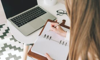 A woman writes in a notebook while using a laptop to job hunt and create a resume for blog most how to explain employment gaps