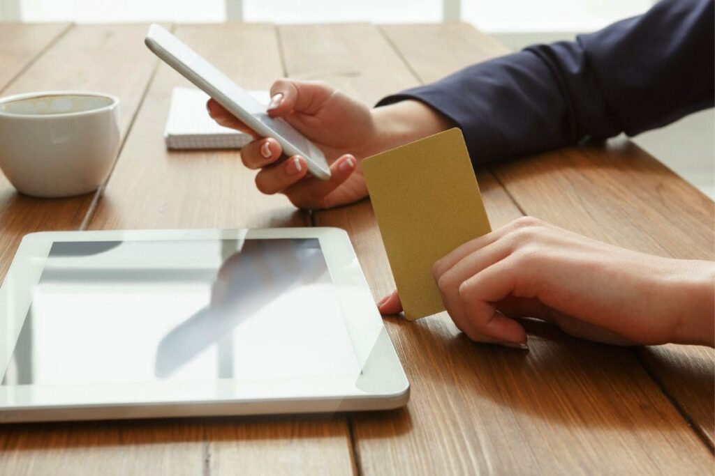 A woman sitting at a cafe, drinking coffee, holding a credit card, and using her phone to shop online.