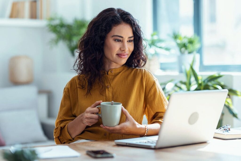 A woman working from her home office desk, holding a coffee cup, smiling, and reading on her laptop.