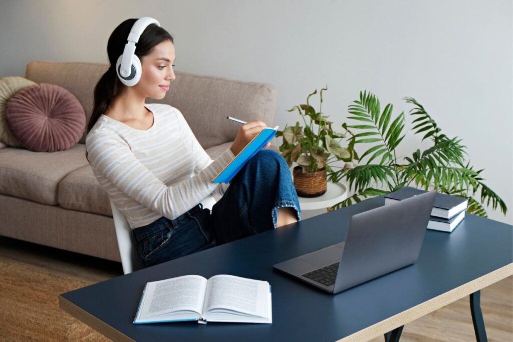 A woman working from home at a desk, writing in a notebook and using a laptop.