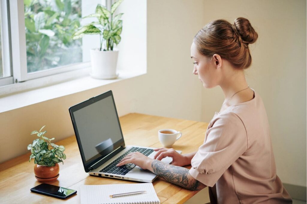 A woman working from her home office desk, using a laptop and writing in a notebook.