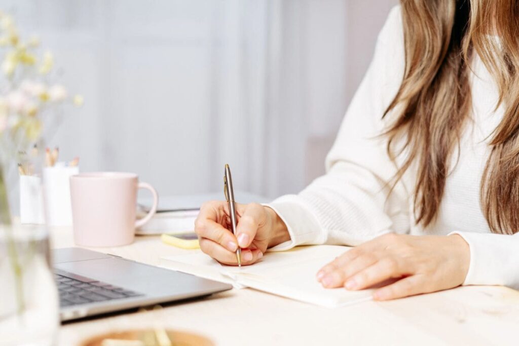 Una mujer que trabaja desde el escritorio de su oficina en casa, usando una computadora portátil y escribiendo en un cuaderno.