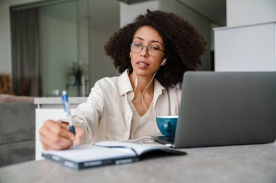A woman working from home at her kitchen table, using a laptop and writing in a notebook for the blog post how to get a remote job.