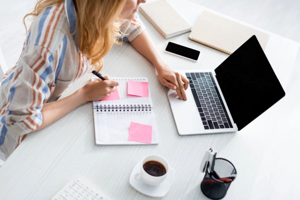 Una mujer que trabaja desde el escritorio de su oficina en casa, usando una computadora portátil y escribiendo en un cuaderno.
