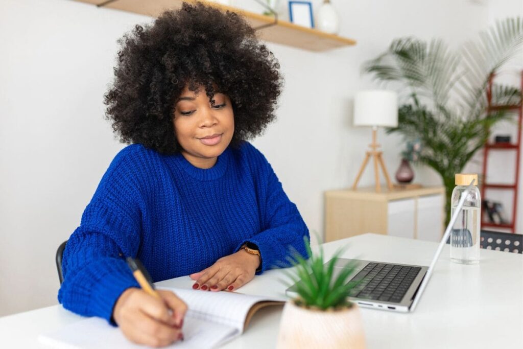A woman working from home in a modern office, using a laptop and writing in a notebook.