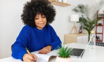 A woman working from home in a modern office, using a laptop and writing in a notebook.